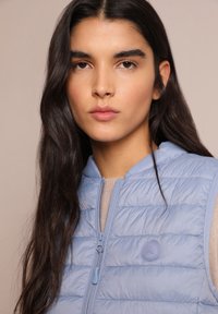 Gilet trapuntato blu chiaro con zip frontale e cappuccio. Presenta una toppa con logo e una texture liscia e lucida. Indossato sopra una maglia beige.