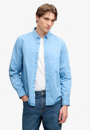 Ung mand med lysebrunt hår iført en lyseblå skjorte over en hvid t-shirt og blå jeans, poserer med den ene hånd i lommen.
