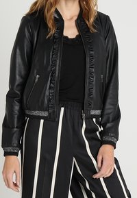 Žena v černé bundě z umělé kůže s volánkovým lemem a pruhovanými černobílými kalhotami s vysokým pasem.