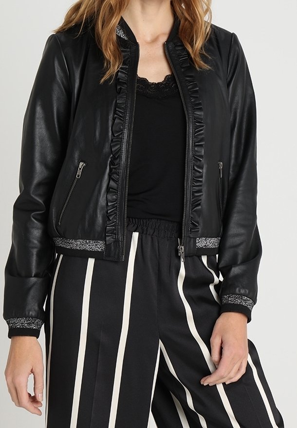 Žena v černé bundě z umělé kůže s volánkovým lemem a pruhovanými černobílými kalhotami s vysokým pasem.