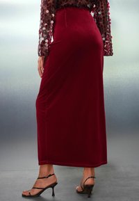 Falda larga de terciopelo rojo con forma ajustada, abertura lateral y textura suave. Llevada con zapatos de tacón negro con tiras delgadas y tacón de aguja.