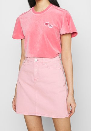 Femme portant un t-shirt en velours rose avec un cœur et le logo Adidas, associé à une jupe en denim taille haute rose clair.