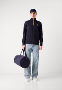Hanorac bleumarin cu fermoar galben, patch cu logo; blugi deschiși, cu talie largă; geantă de voiaj bleumarin; adidași albi; șapcă de baseball.
