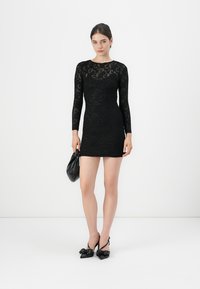 Robe longue à manches longues en dentelle noire avec une silhouette ajustée. Texture lisse, décolleté rond et longueur mini. Portée avec des accessoires noirs.