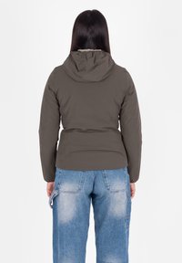 Vrouw in een bruine gewatteerde jas met capuchon en loszittende blauwe spijkerbroek, staand met de rug naar de camera tegen een witte achtergrond.