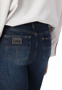 Mörkblå jeans med en åtsittande silhuett. Har två bakfickor med en svart etikett som säger "Lois." Sömnaden har en kontrasterande gul färg.