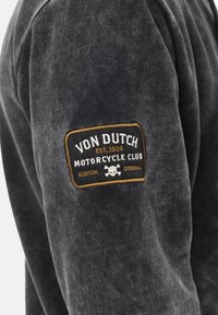 Sweatshirt noir en tissu doux avec un patch texturé. Le patch comprend "Von Dutch Motorcycle Club", un graphique de crâne et des accents dorés.