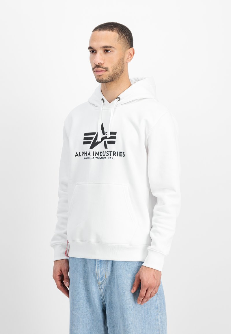 Sudadera blanca con un bolsillo frontal, que presenta un logo negro y el texto "ALPHA INDUSTRIES". Tejido texturizado, cordones y puños acanalados.