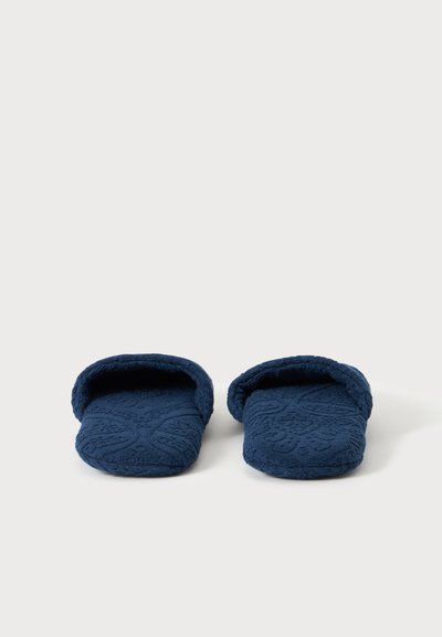 Paire de chaussons bleu foncé texturés à dos ouvert, placés côte à côte sur une surface blanche.