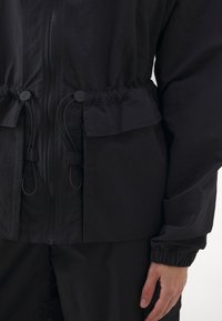 Sort letvægtsjakke med lynlås, justerbare snore i taljen og indsamlede manchetter. Har to frontlommer.