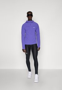 Lila, långärmad, huva idrottströja med en framficka med dragkedja; kombinerad med svarta tights med en logotyp. Modellen bär vita sneakers och solglasögon.