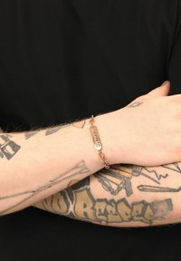 Pulsera de oro rosa con una placa rectangular alargada y un diseño de cadena enlazada, llevada en una muñeca tatuada sobre un fondo negro.