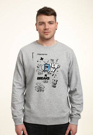 Młody mężczyzna w szarej bluzie z kolorowym kreskówkowym telewizorem i tekstem w stylu graffiti, zawierającym "Love TV", "BREAKS" i "Spin".