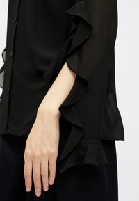 Blusa preta transparente com mangas franzidas e detalhe de botões. O tecido é suave, com uma textura lisa e um corte descontraído.