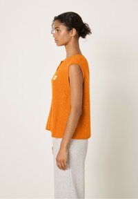 Oranje gebreide mouwloze top met een gestructureerde afwerking en een knoopsluiting aan de voorkant, gedragen over grijze joggingbroeken.