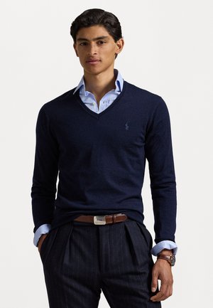 Maglione a v blu navy in morbido tessuto, indossato sopra una camicia colletto azzurro chiaro. Presenta un piccolo logo sul petto ed è abbinato a pantaloni con righe sottili.