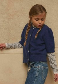 Ragazza con i capelli intrecciati che indossa un cardigan blu navy sopra una camicia bianca a maniche lunghe in pizzo e jeans blu, appoggiata a una parete beige con texture.