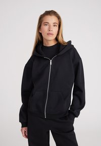 Sudadera negra con cremallera fabricada en material suave, que cuenta con una capucha, bolsillos frontales y puños acanalados. El diseño es de corte holgado.