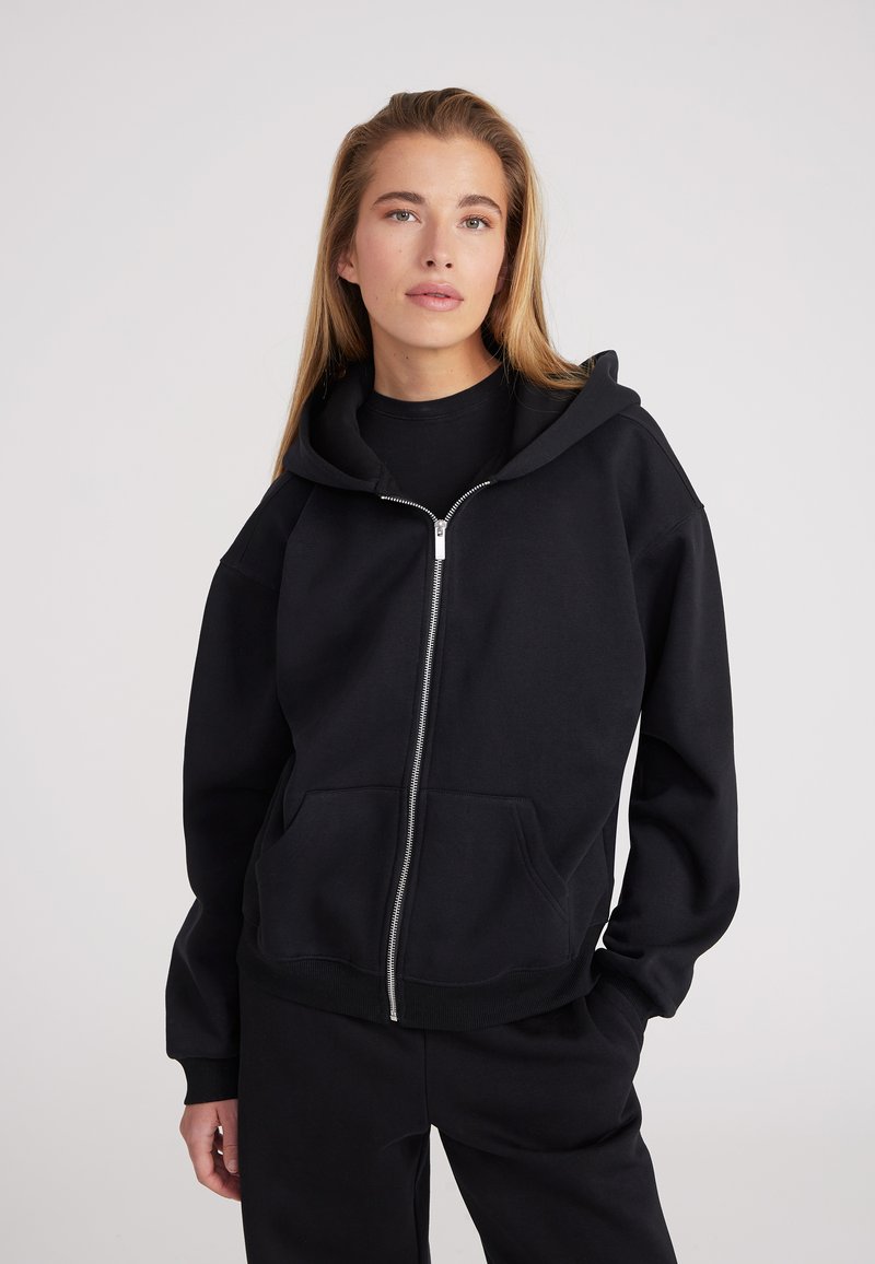 Sudadera negra con cremallera fabricada en material suave, que cuenta con una capucha, bolsillos frontales y puños acanalados. El diseño es de corte holgado.