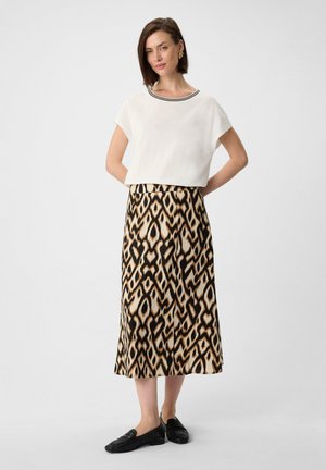 Witte crop top met zwarte halslijn, gecombineerd met een lange rok met een gedurfde geometrische print in zwart, beige en bruin. Zwarte platte schoenen.