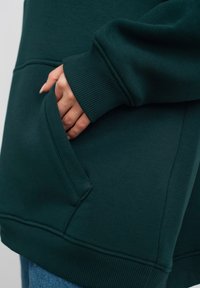 Donkergroene hoodie met een kangoeroezak, ribbelmanchetten en een zachte textuur. Close-up van een hand die in de zak steekt.