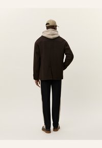 Chaqueta marrón de tela texturizada, sudadera con capucha beige debajo, pantalones negros con franjas laterales y zapatos marrones claros.