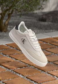 Sneaker bej Calvin Klein cu talpă și sireturi albe, detaliu pe tocul negru și logo-ul "ck", plutind deasupra plăcilor pătrate din lemn și a unor pietricele mici.