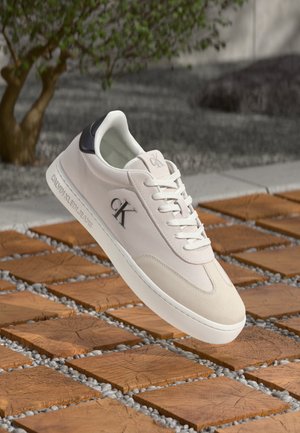 Beige Calvin Klein sneaker with white sole and laces, black heel detail, and "ck" logo, floating above wooden square tiles and small pebbles.