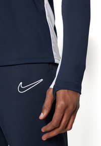 Nærbillede af en person iført navyblå og hvide træningsklæder med et hvidt Nike-logo på bukserne og et tommelfingerhul på ærmet.