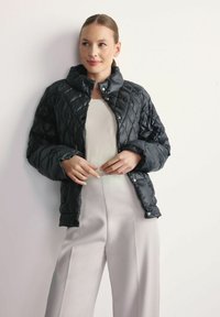 Pikowana czarna kurtka puchowa z wysokim kołnierzem i zapięciem na zatrzaski, noszona na jasnej bluzce w zestawieniu z kremowymi spodniami o szerokich nogawkach.