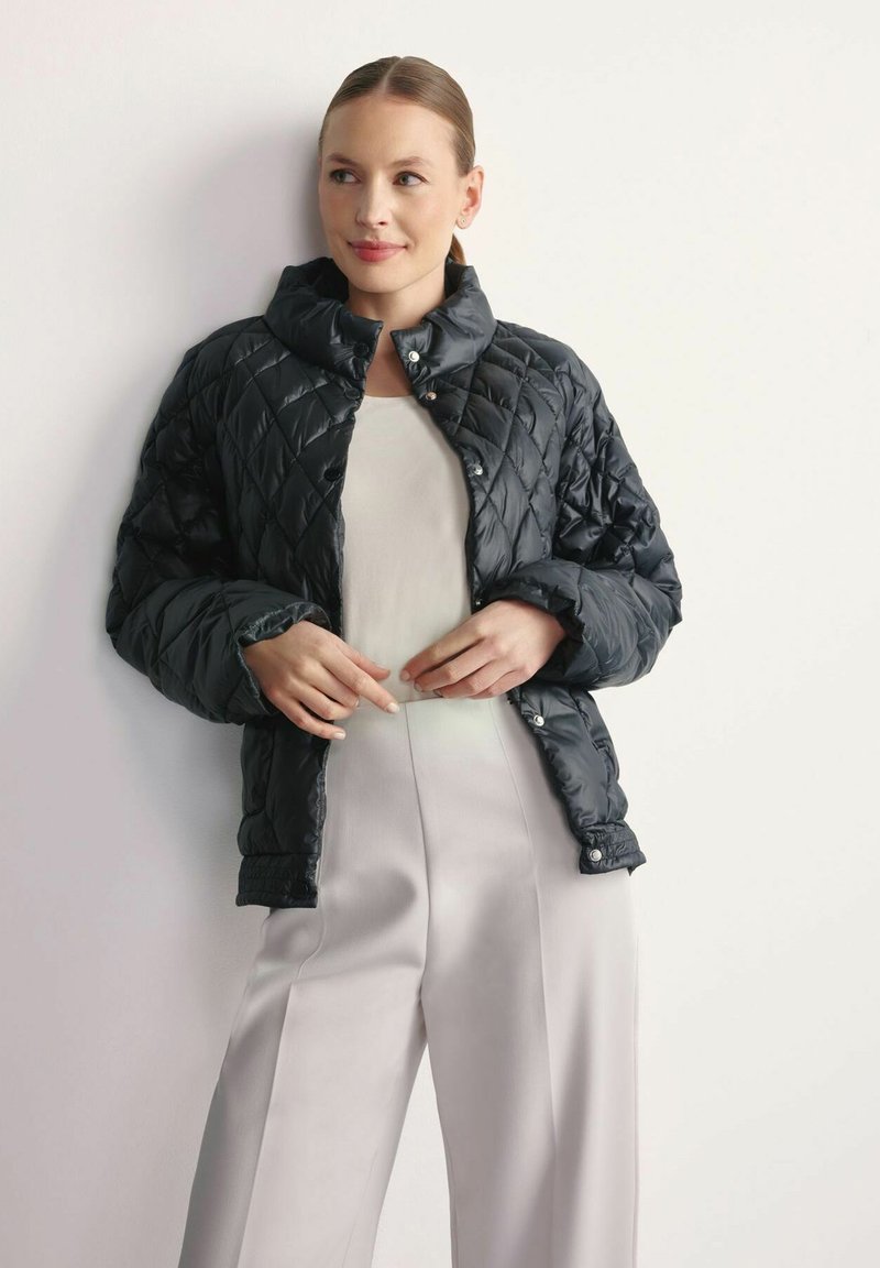 Pikowana czarna kurtka puchowa z wysokim kołnierzem i zapięciem na zatrzaski, noszona na jasnej bluzce w zestawieniu z kremowymi spodniami o szerokich nogawkach.