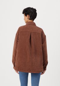 Camicia oversize in corduroy marrone con una piega sul retro, spalle cadenti e polsini con bottoni, abbinata a jeans in denim blu.