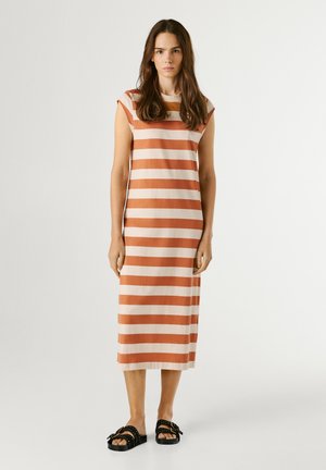 Žena stojaca v nosí midi šaty bez rukávov, s oranžovo béžovými horizontálnymi pásikmi a čierne slip-on sandále s prackami.