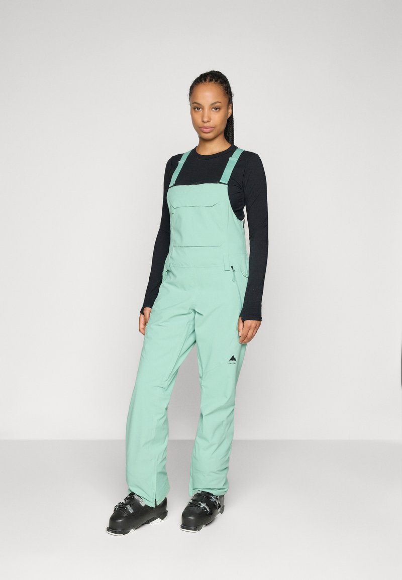 Overoles de esquí verde menta con tirantes ajustables, bolsillos frontales y piernas ajustadas, combinados con una camiseta de manga larga negra y botas de esquí negras.