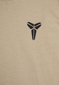 Beige pamut póló, bal mellen fekete geometrikus logóval. Az anyag sima textúrájú, kerek nyakkivágással.