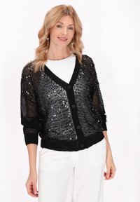 Svart, paljetterad cardigan med en lös stickad design. Har en V-hals, knappstängning och ribbade ärmslut. Bärs över en vit skjorta.