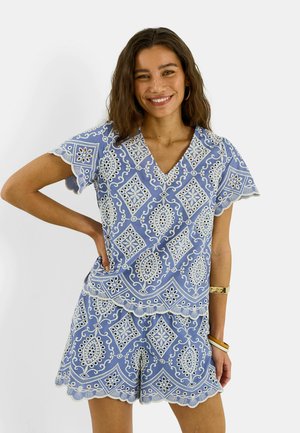 Mujer sonriendo con la mano en la cadera, vistiendo un top azul con estampado en forma de V y pantalones cortos a juego con bordes ondulados sobre fondo blanco.