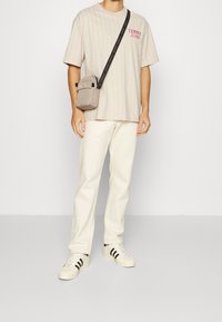 Beige stribet t-shirt med rød logo, cremefarvede bukser, sorte-stribede hvide sneakers og en grå crossbody taske med justerbar rem.