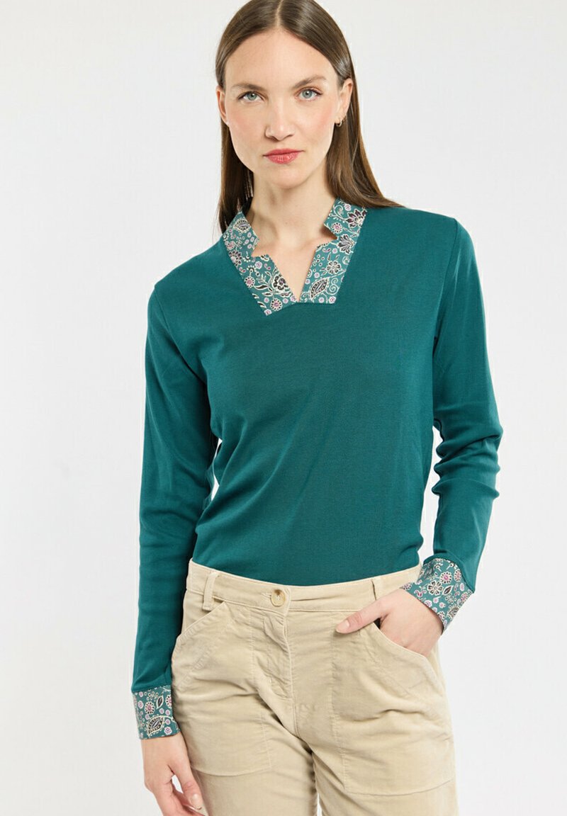 T-shirt à manches longues turquoise avec un col en V et des poignets à motifs. Le tissu est lisse et le design a une coupe décontractée.