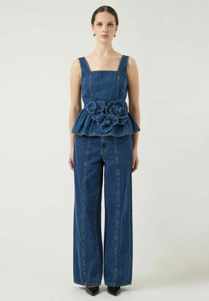 Donna con top smanicato in denim blu scuro con dettagli floreali 3D e jeans a gamba larga abbinati, in piedi davanti a uno sfondo chiaro e uniforme.