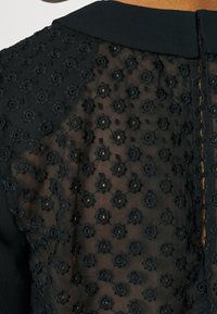 Blusa nera con un retro trasparente e ricamato, caratterizzato da motivi floreali e piccole aperture, completata da un colletto nero uniforme.