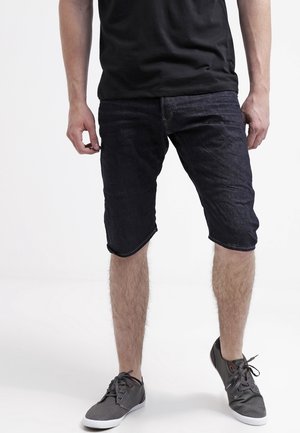 Homme portant un t-shirt noir, un short en jean foncé et des baskets grises à lacets, debout sur fond blanc.