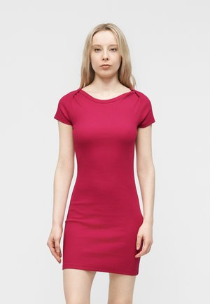 Giovane donna con lunghi capelli biondi che indossa un vestito aderente color magenta a maniche corte, in piedi contro uno sfondo grigio chiaro uniforme.