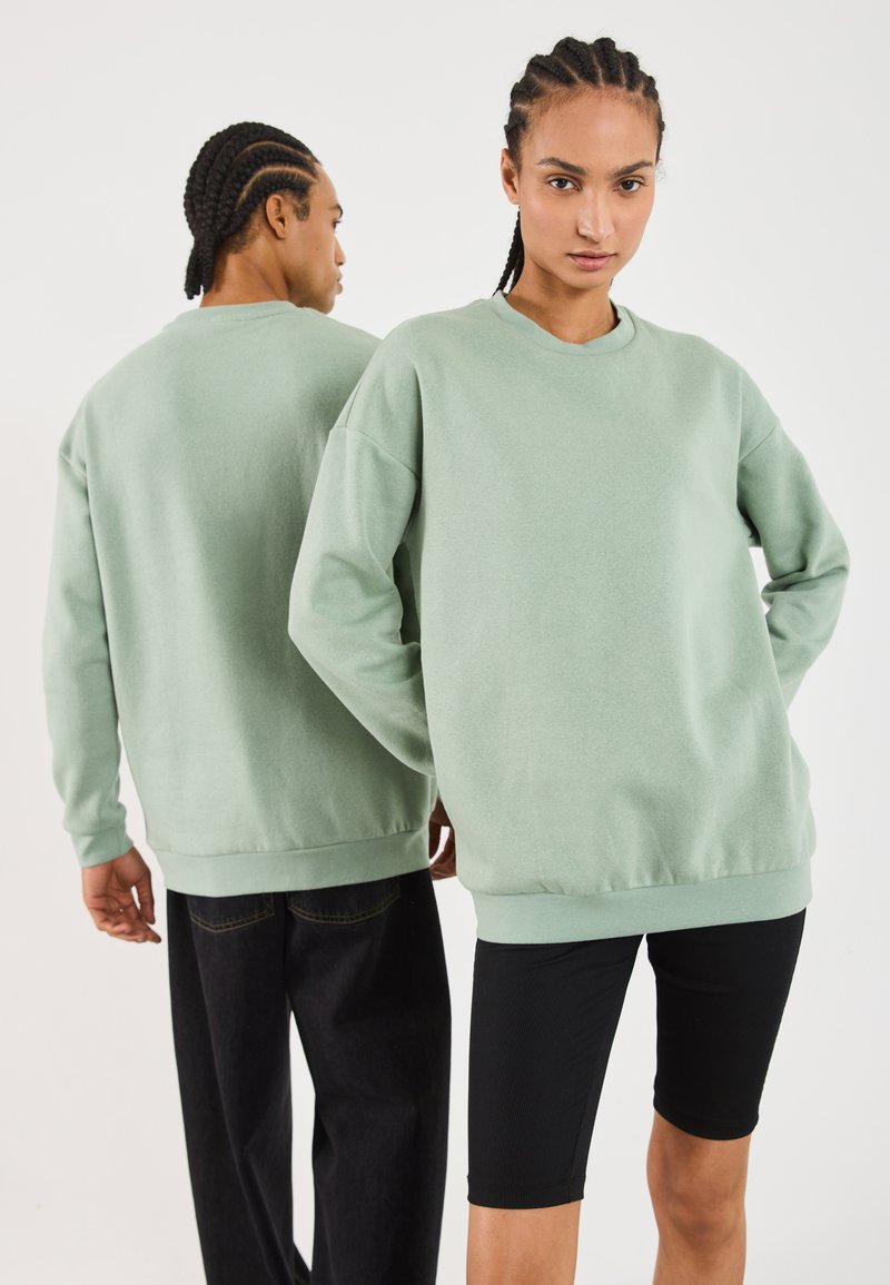 Dos modelos con cabello trenzado que llevan sudaderas grandes de color verde claro a juego, uno de frente y el otro de espaldas.