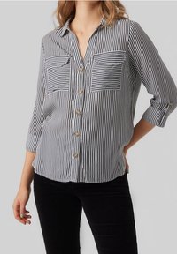 Chemisier à rayures noires et blanches avec col, manches courtes, présentant deux poches poitrine et des boutons en laiton sur le devant. Texture de tissu douce.