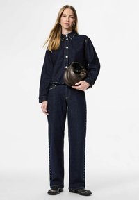 Mørk denimjakke med knappelukking og nagledetaljer, kombinert med matchende vidbukser med nagler langs sidene. Holder en brun veske.