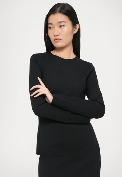 Fekete, hosszú ujjú felső sima anyagból, kerek nyakkivágással és testhezálló szabással. A modell karját keresztbe téve pózol világos háttér előtt.