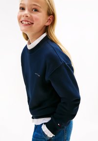 Felpa blu navy realizzata in tessuto morbido, con colletto e polsini a costine, presenta un piccolo logo sul petto. Abbinata a una camicia bianca sotto.