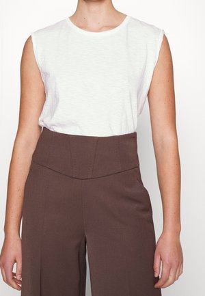 Osoba nosząca bezrękawnik w białym kolorze, włożony w wysokie brązowe spodnie na jednolitym białym tle.