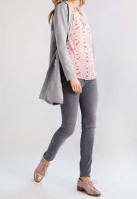 Cardigan long gris sur une blouse rose à motifs, jean slim gris et chaussures à lacets dorées rose brillant. Tissu lisse avec une texture subtile.